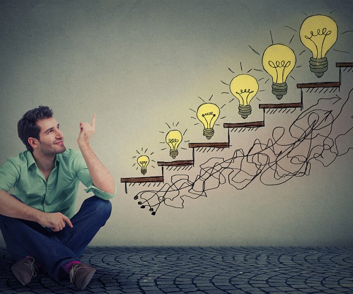 Happy man sitting on floor in his office pointing at business education success, promotion, company growth isolated gray wall texture background. Handsome guy looking up at growing up light bulbs